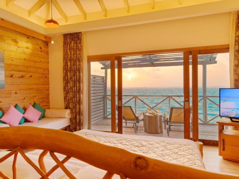 Chambre d'hôtel avec plafond en bois, lit confortable, canapé et vue sur la mer au coucher du soleil.