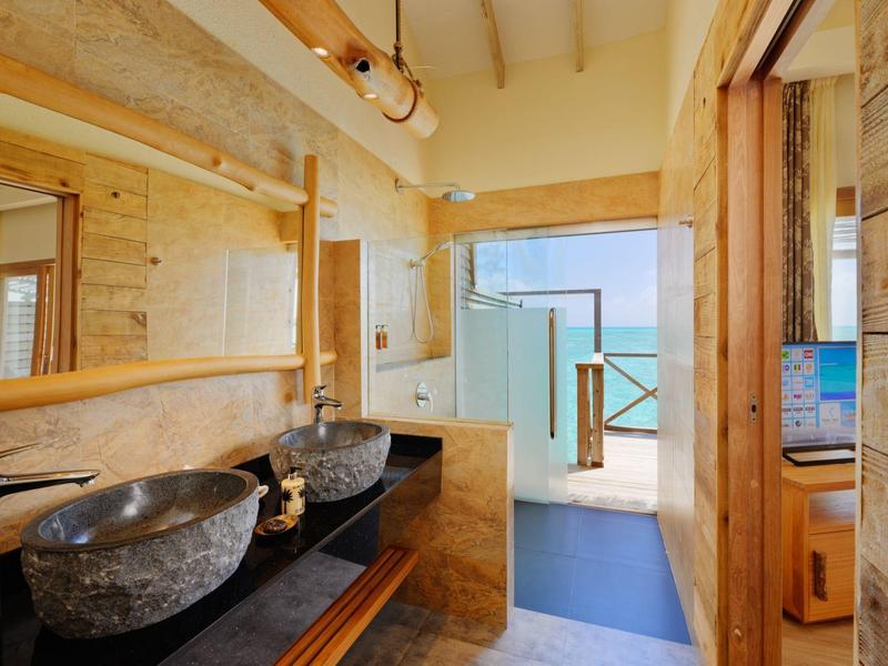 Salle de bain lumineuse avec lavabos en pierre, détails en bois et vue sur la mer par la porte ouverte.