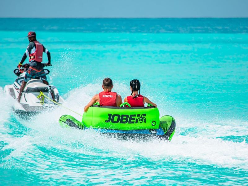 Deux personnes sur un bateau gonflable vert de sport nautique derrière un jet-ski sur une eau turquoise.