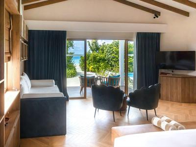 Chambre d'hôtel lumineuse avec poutres en bois, coin salon et vue sur terrasse avec jardin vert et mer.