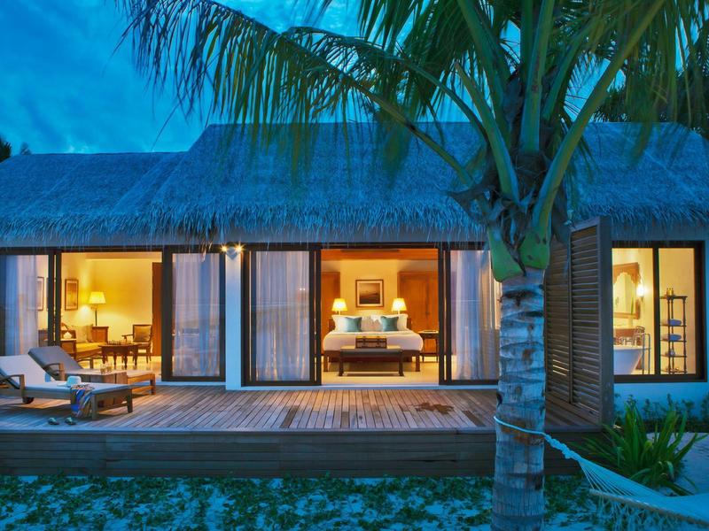 Villa moderna sulla spiaggia con terrazza in legno, lettini e palme illuminate la sera.