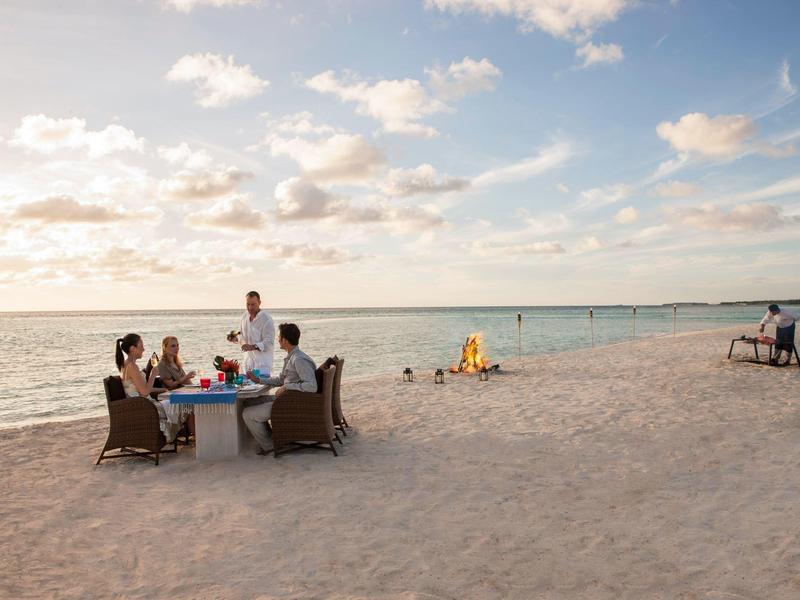 Persone che gustano un pasto a un tavolo apparecchiato sulla spiaggia con vista sul mare.