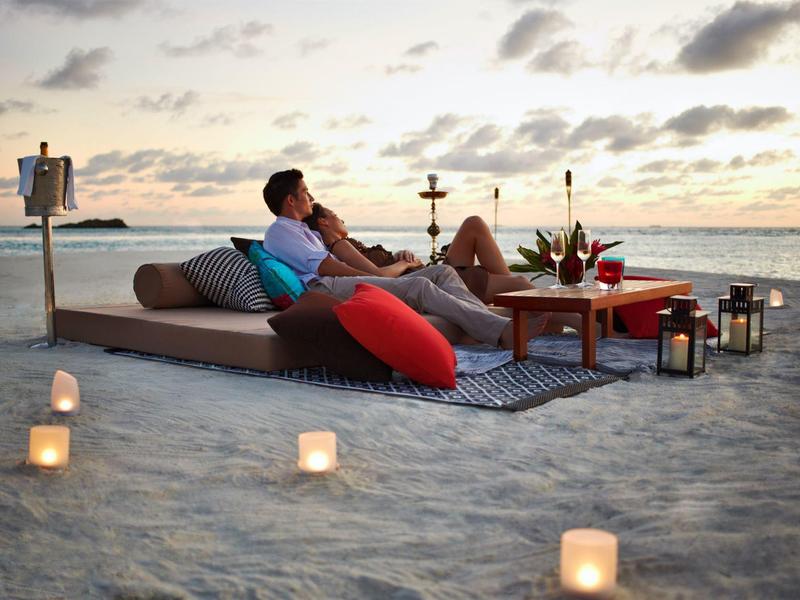 Coppia seduta su un comodo divano in spiaggia al tramonto, circondata da candele e decorazioni.