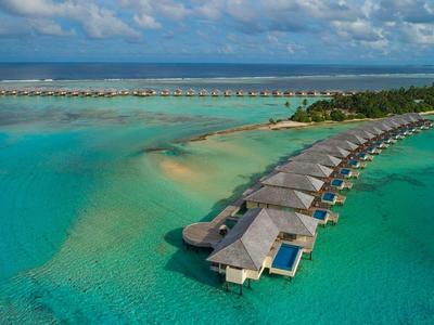 Overwasser-Bungalows an türkisfarbenem Meer mit Strand und Palmen unter blauem Himmel.