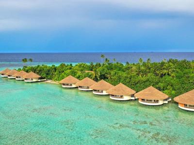 Reihe von Bungalows mit Strohdächern über klarem, türkisfarbenem Wasser vor grüner Inselkulisse.