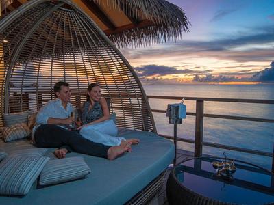 Paar sitzt entspannt auf einer großen, runden Lounge unter Palmen bei Sonnenuntergang am Meer.