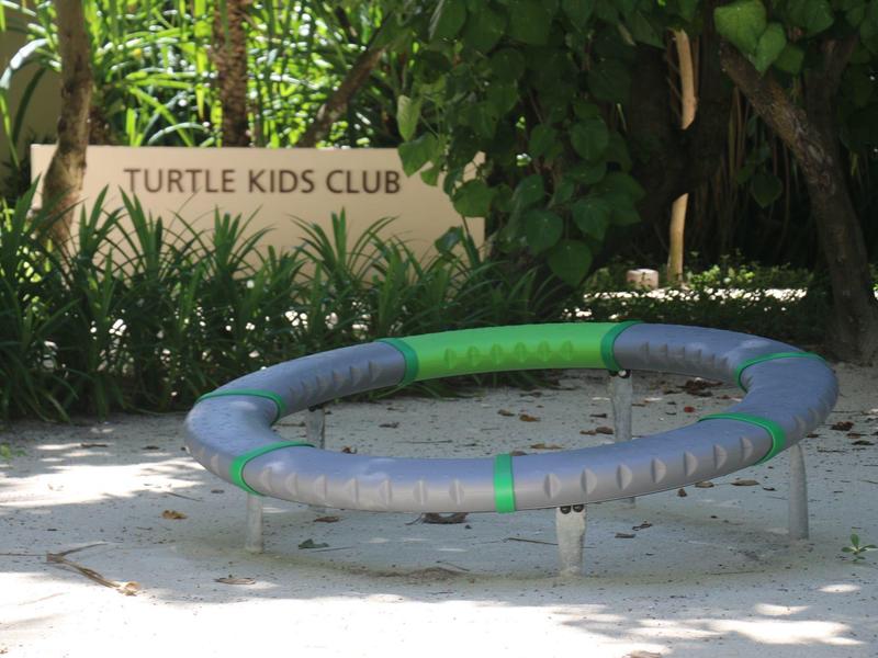 Kleines graugrünes Raumschiffspielgerät auf Sandboden vor grünem Blattwerk und Schild "Turtle Kids Club".