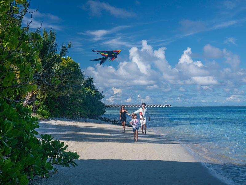 Menschen am Sandstrand neben blauem Meer, Drohne fliegt über ihnen, bewachsene grüne Bäume links.