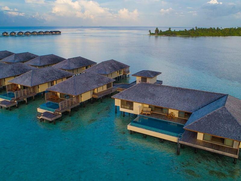 Überwasserbungalows mit Strohdächern auf türkisfarbenem Wasser, im Hintergrund eine kleine Insel und blauer Himmel.