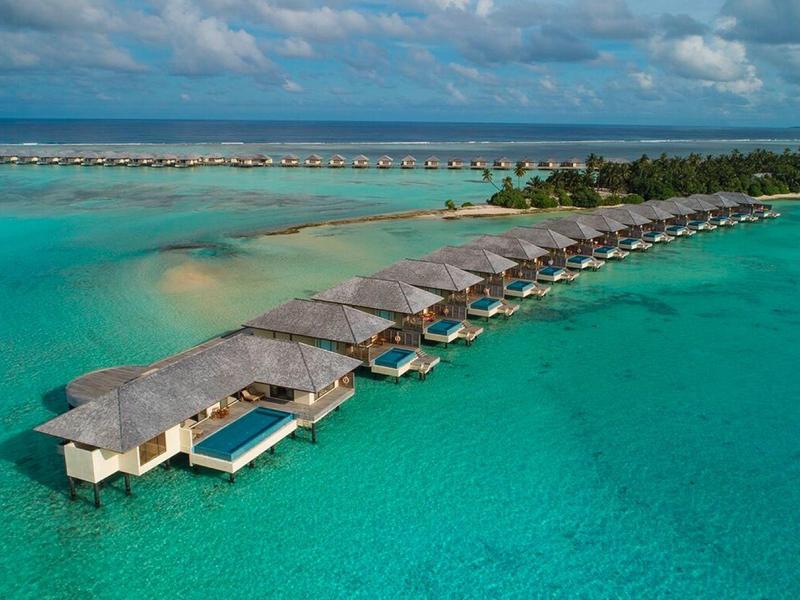 Bungalows mit Strohdächern auf Stelzen im türkisblauen Wasser eines tropischen Meeres.