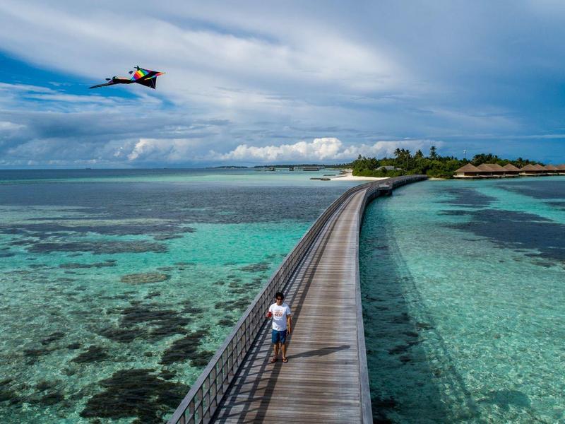 Holzsteg führt über türkisfarbenes Meer zu kleiner Insel unter bewölktem Himmel, Drohne fliegt links.