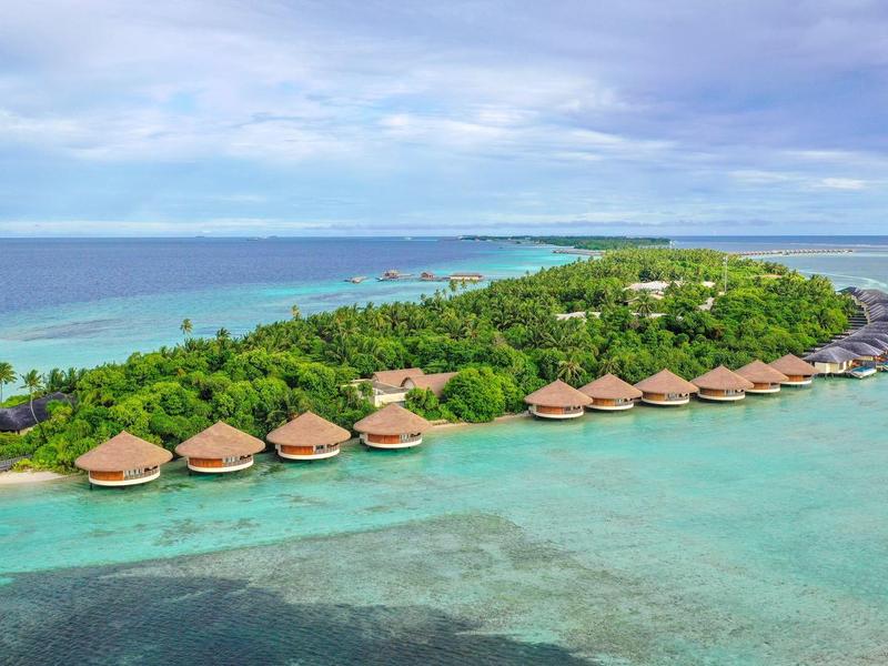 Grüne Insel mit Palmen und Holzhütten am türkisblauen Meer unter bewölktem Himmel.