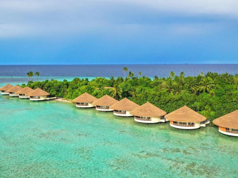 Reihe von Bungalows mit Strohdächern über klarem, türkisfarbenem Wasser vor grüner Inselkulisse.