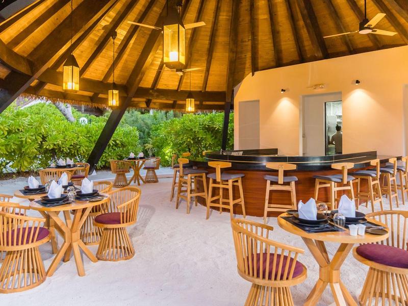 Offener, runder Barbereich mit Holzmöbeln und Strohdach, umgeben von grüner Vegetation.