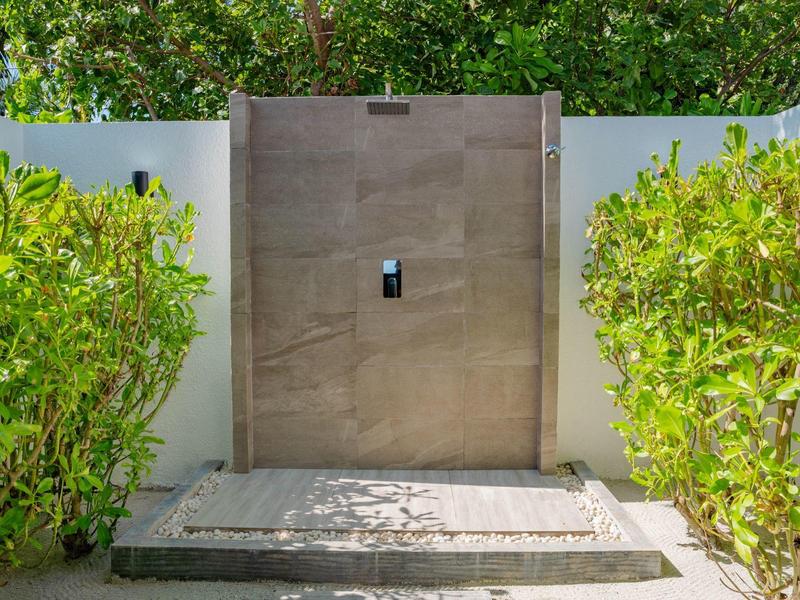 Moderner Eingang mit brauner Holztür, weißer Mauer und grünen Sträuchern links und rechts.