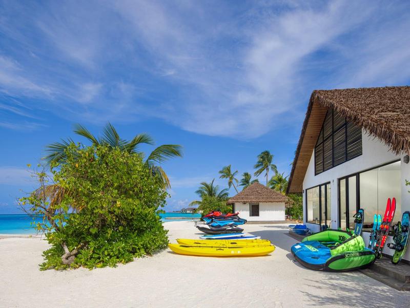 Weißer Sandstrand mit bunten Kajaks, Palmen und einem Haus mit Strohdach unter blauem Himmel.
