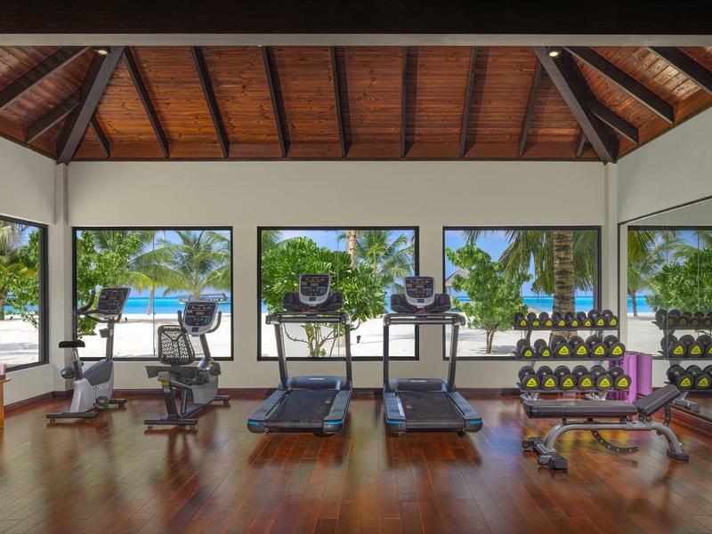 Moderner Fitnessraum mit Holzboden, drei Laufbändern, Hanteln und großen Fenstern mit Meerblick.