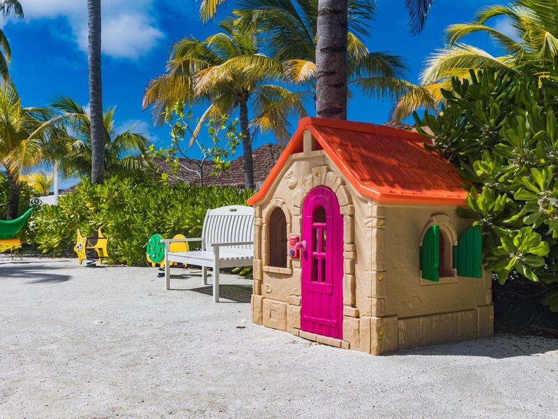 Kleines Spielhaus aus Kunststoff mit rotem Dach und pinker Tür auf sandigem Boden unter Palmen.