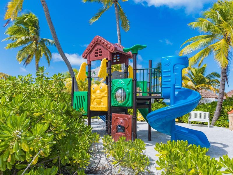 Bunter Spielplatz mit Rutsche, Klettergerüst und Palmen unter blauem Himmel im Sand.