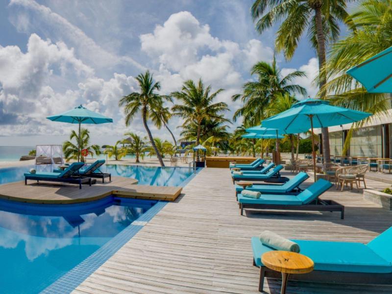 Holzsteg entlang Pool mit blauen Liegestühlen und Sonnenschirmen, Palmen und Himmel im Hintergrund.