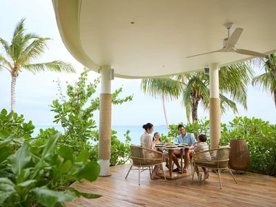 Drie personen zitten aan een tafel onder een overdekt terras met palmbomen.