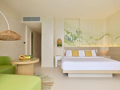 Helles Hotelzimmer mit modernem Bett, grünem Sofa und Wandkunst mit Pflanzenmotiven.
