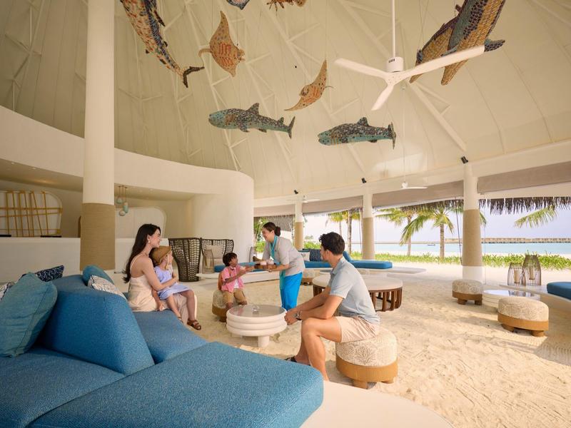 Mensen zitten in een open loungegedeelte aan het strand met een zanderige vloer en vissen aan het plafond.