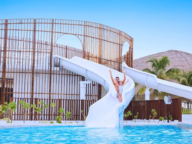 Persoon glijdt van een witte waterglijbaan in een zwembad met modern gebouw en bergen op de achtergrond.