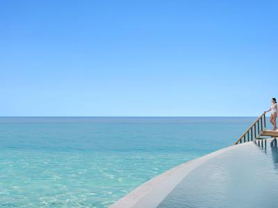Persona sentada al borde de una piscina infinita con vista al mar azul y cielo despejado.