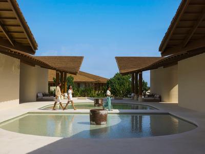 Dos personas sentadas junto a una piscina entre edificios modernos abiertos bajo un cielo despejado.