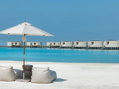 Playa de arena blanca con sombrilla y cojines frente a bungalows sobre el agua en el mar azul cristalino.