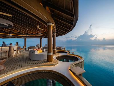 Una terraza con sofás y una pequeña piscina sobre el agua al atardecer.