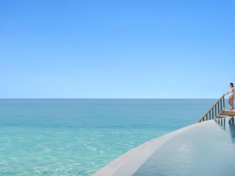 Persona sentada al borde de una piscina infinita con vista al mar azul y cielo despejado.