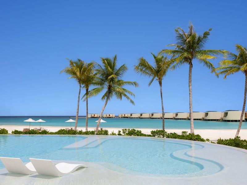Vista de una piscina con tumbonas, palmeras y playa bajo un cielo azul.