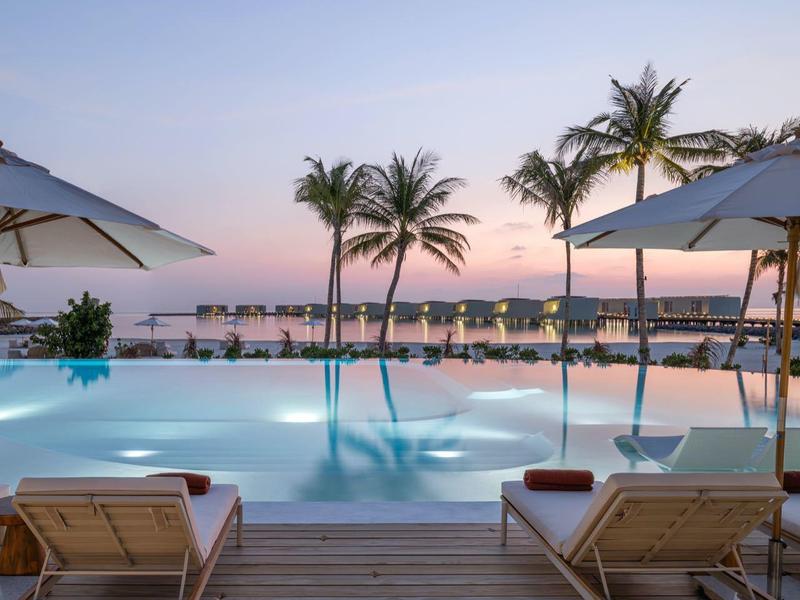 Piscina infinity con tumbonas y sombrillas frente a palmeras al atardecer