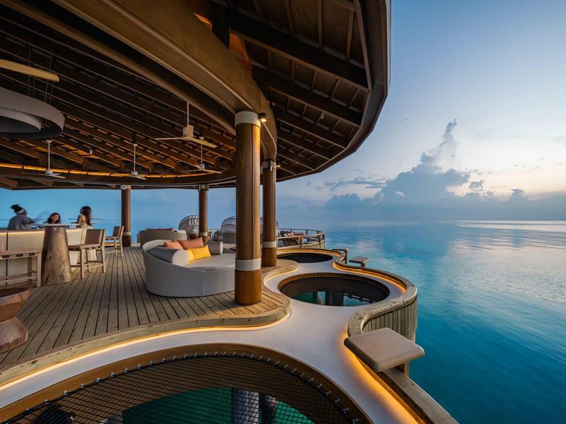 Una terraza con sofás y una pequeña piscina sobre el agua al atardecer.
