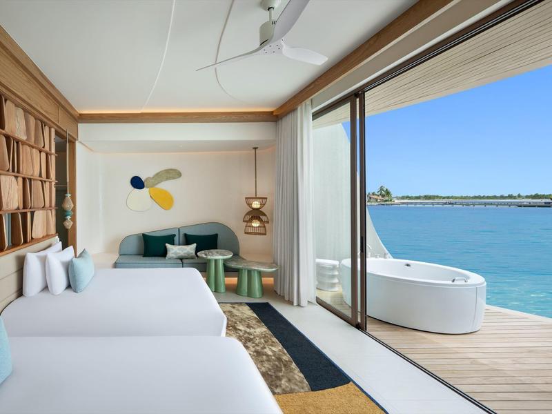 Habitación de hotel moderna con dos camas, sofá, estantería y terraza con jacuzzi con vistas al mar.