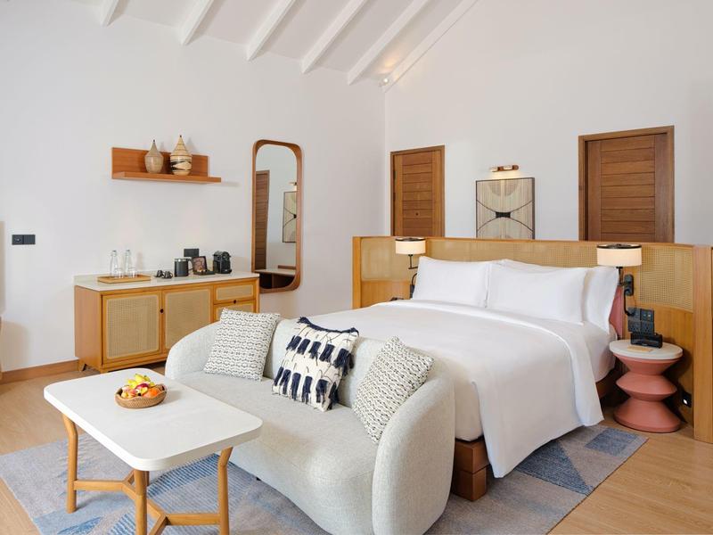 Chambre d'hôtel moderne et lumineuse avec grand lit, canapé, TV et coin salon aux tons chauds.