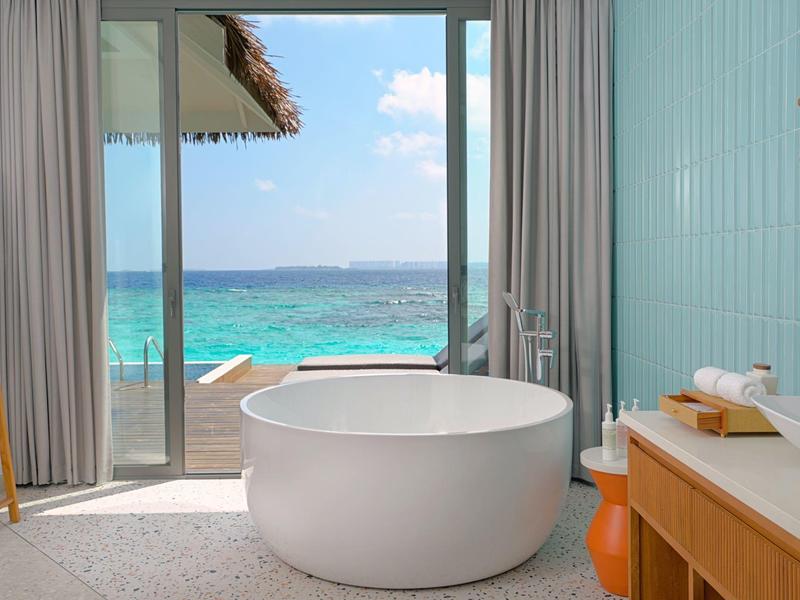 Baignoire blanche autoportante avec vue sur la mer bleue et le ciel à travers une grande porte coulissante en verre.