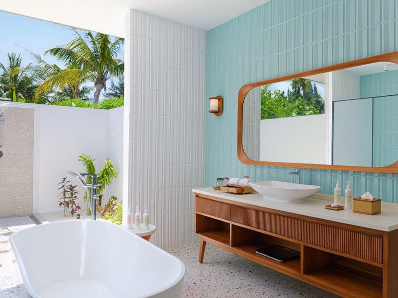 Salle de bain lumineuse avec baignoire autoportante, grand meuble en bois et douche extérieure dans un complexe tropical.