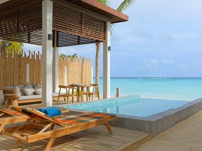 Blick auf einen Pool mit Liegestühlen und Holzmöbeln direkt am Strand bei klarem Himmel.