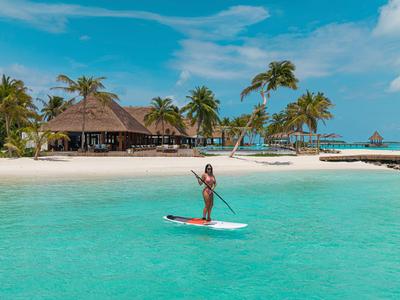 Frau auf einem Paddleboard im klaren türkisfarbenen Wasser vor einer tropischen Strandbar mit Palmen.