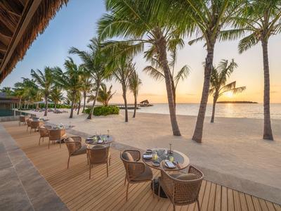 Area pranzo all'aperto sulla spiaggia con palme e vista sul tramonto sul mare.