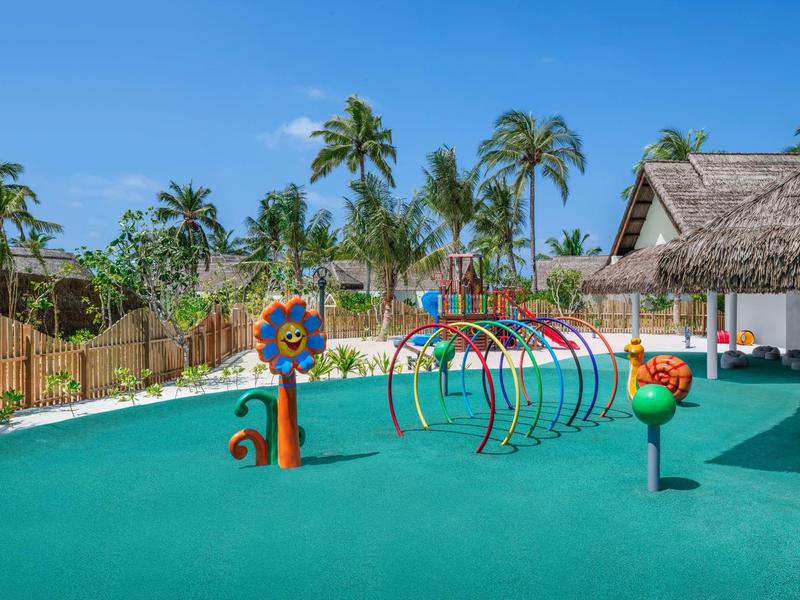 Parco giochi colorato per bambini con attrezzature sotto palme e cielo soleggiato.