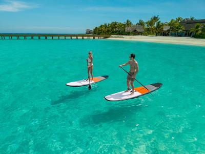 Dos personas reman en tablas de paddle surf sobre agua turquesa clara cerca de una isla tropical.