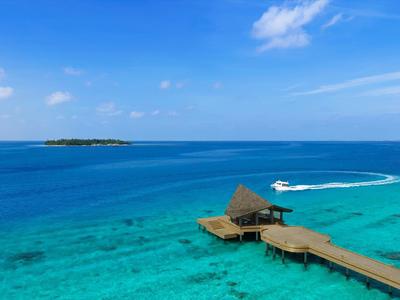 Hölzerner Steg führt zu einem über dem türkisfarbenen Wasser stehenden Strohdachpavillon unter blauem Himmel.