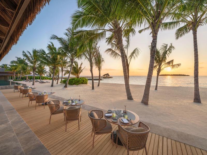 Strandrestaurant mit Tischen und Stühlen unter Palmen bei Sonnenuntergang am Meer.