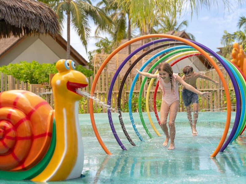 Kinder spielen in einem Wasserpark mit bunten Bögen und einer Schneckenskulptur.