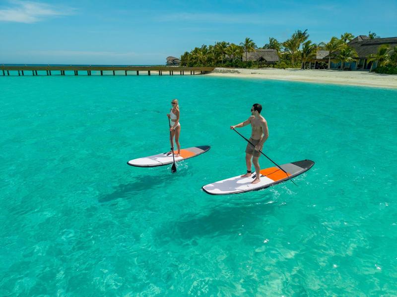 Zwei Personen paddeln auf Stand-Up-Paddle-Boards in klarem, türkisfarbenem Wasser vor einer tropischen Insel.
