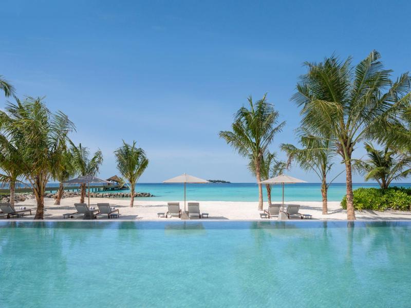 Ein Pool mit Palmen am Strand und Blick auf das blaue Meer unter klarem Himmel.
